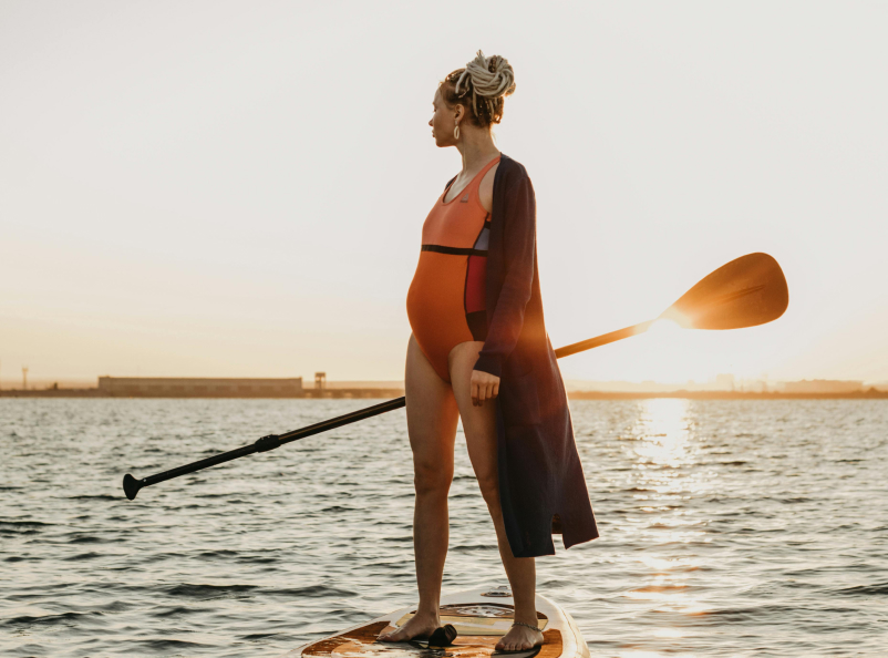 A pregnant woman on a stand up kayack staring over her shoulder at the horizon
